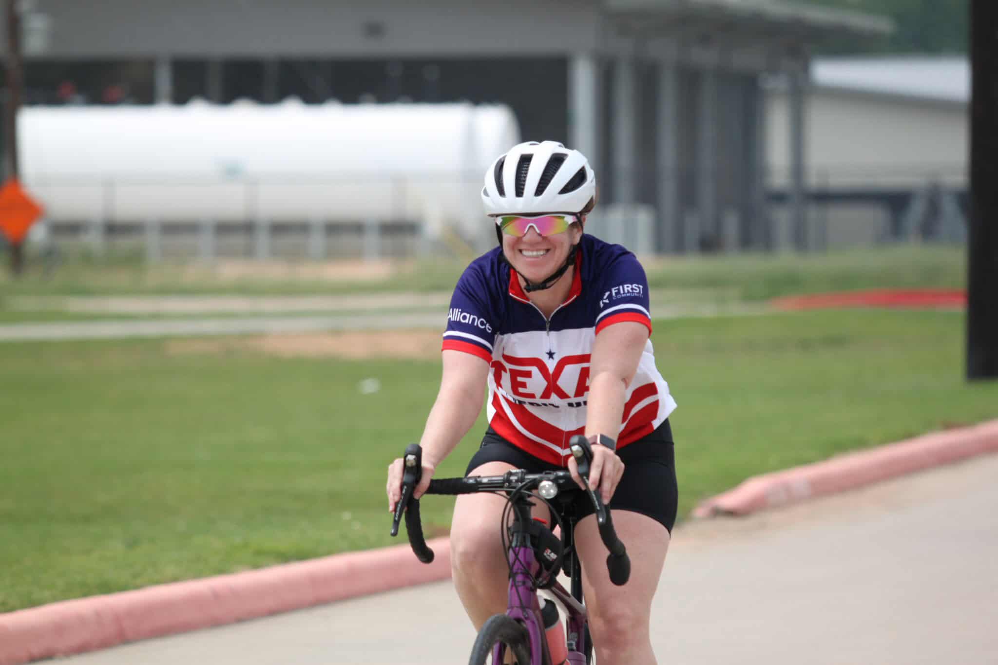 Bright-eyed woman riding a bike wearing a Dallas Texans jersey, white helmet, and sunglasses on a sunny day.