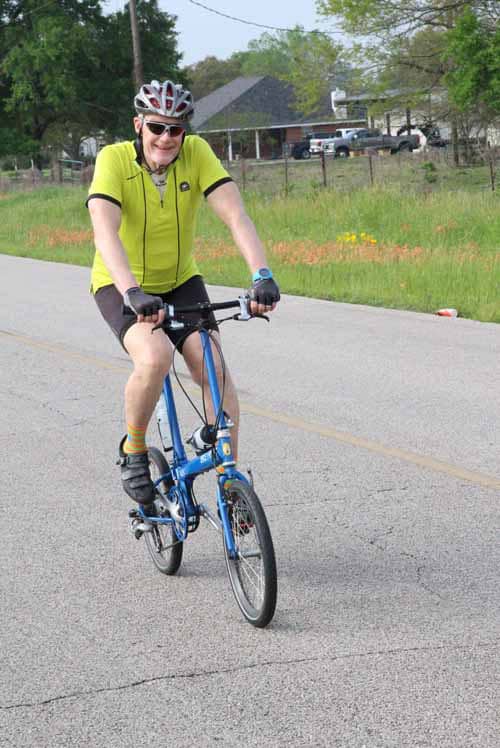 Smiling cyclist riding a blue bike on a suburban road with greenery in the background.