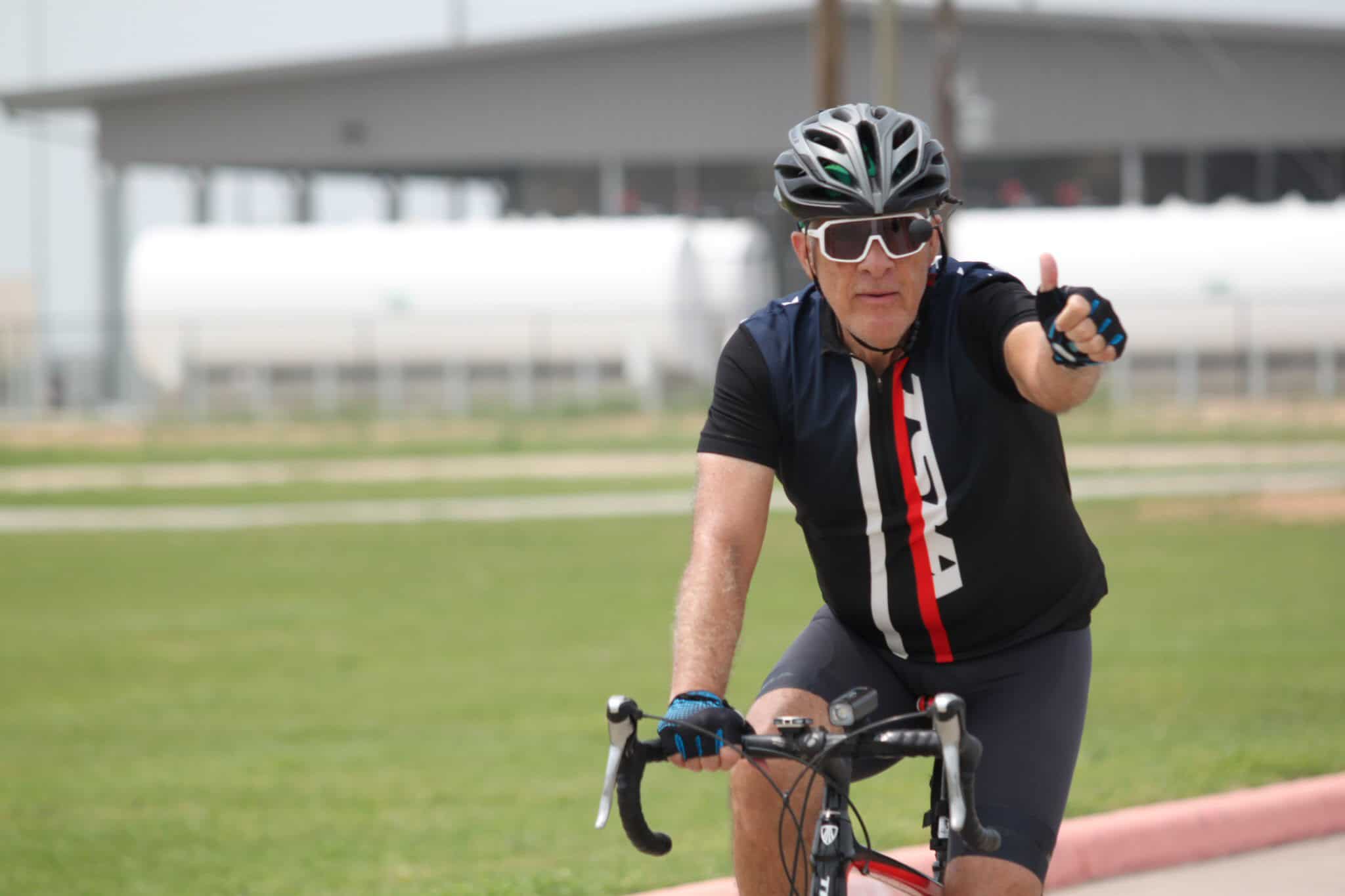 Older male cyclist wearing a helmet and sunglasses, giving a thumbs-up during outdoor ride for Bluebonnet Express.