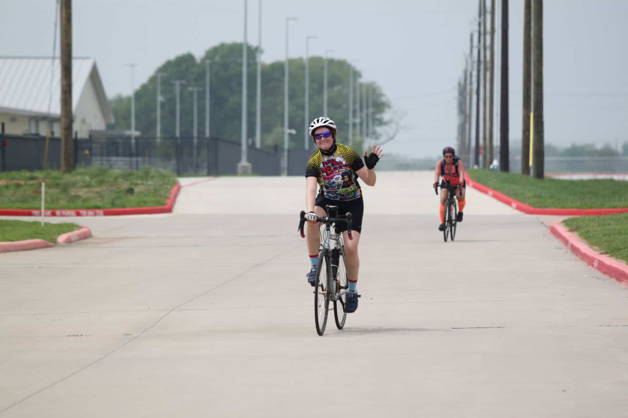 Smiling woman riding a bicycle on a paved path, waving, with fellow cyclist in the background.
