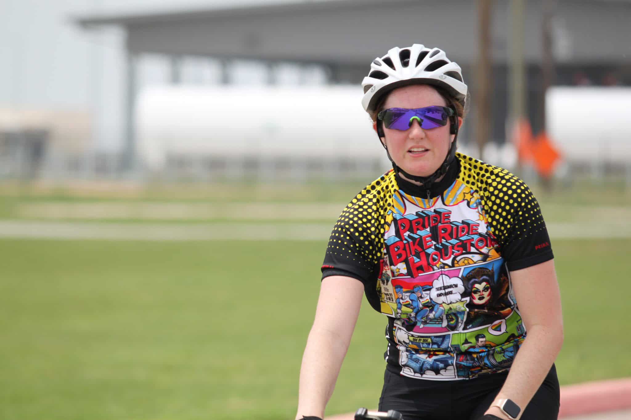 Bright female cyclist wearing Houston Pride Bike Ride jersey, helmet, and sunglasses outdoors.