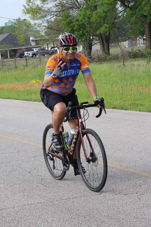 Bright cyclist wearing Bluebonnet Express jersey riding a road bike outdoors.