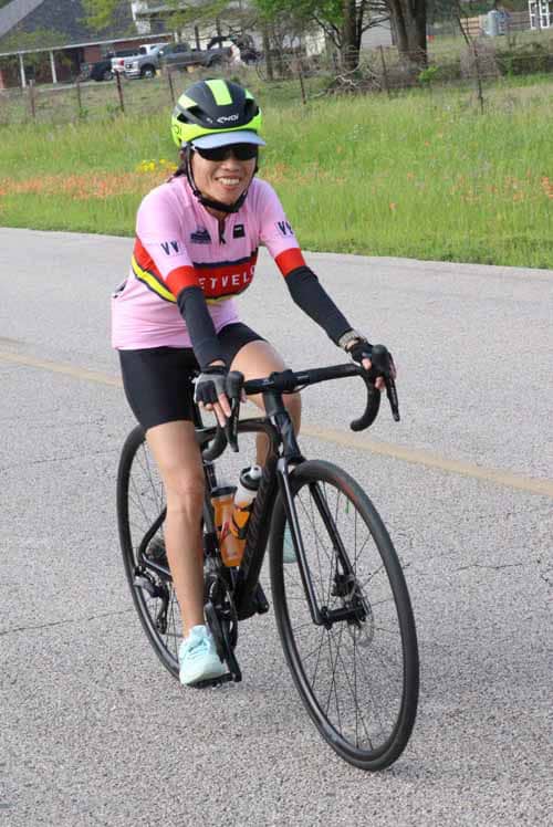 Women cycling on road in pink jersey, helmet, and sunglasses for Bluebonnet Express event.