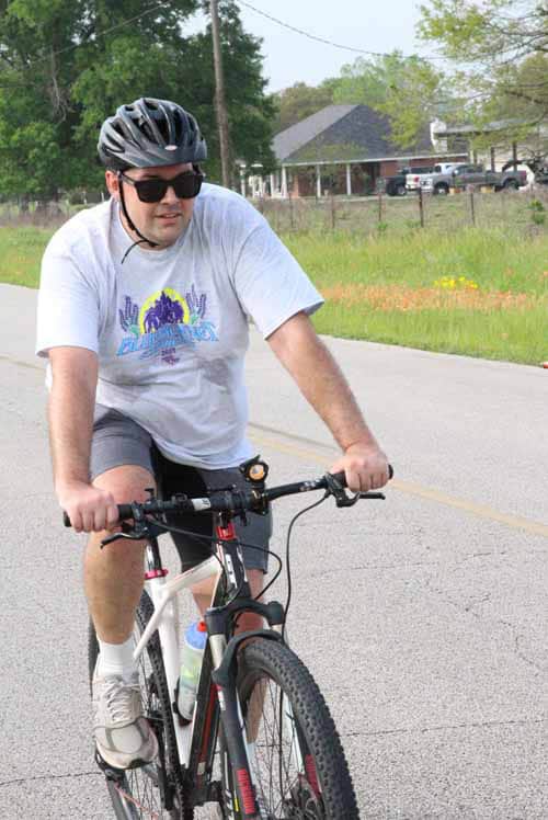A man riding a mountain bike on a rural road with greenery and houses in the background.