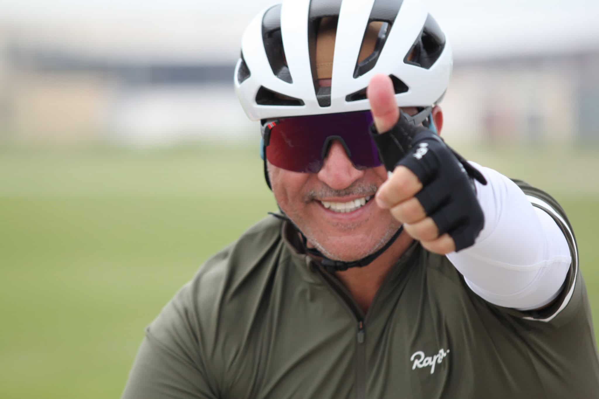 Velo cyclist wearing helmet and sunglasses, giving a thumbs-up outdoors.