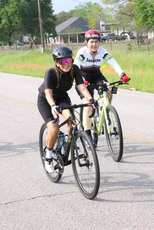 Women riding bicycles on a scenic rural road, promoting cycling events like Bluebonnet Express.