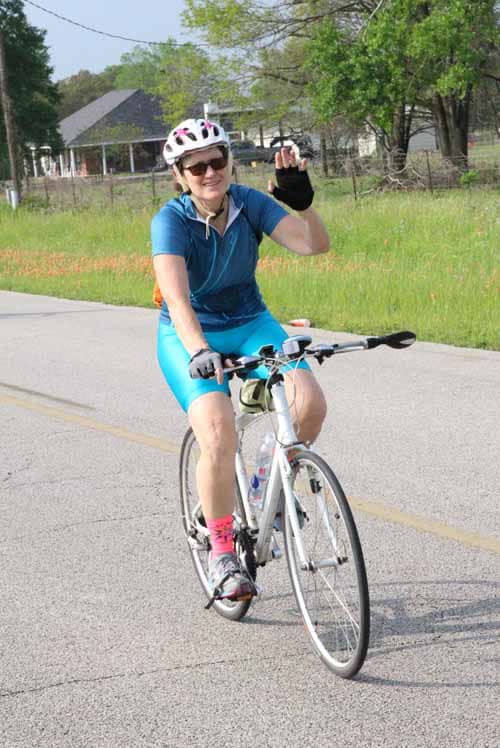 Bright woman riding a bicycle during Bluebonnet Express event, smiling and waving, in a green park setting.