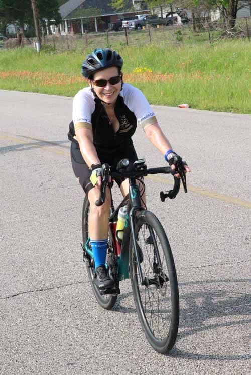 Smiling female cyclist riding a road bike outdoors, wearing a helmet and sunglasses, on a scenic route.
