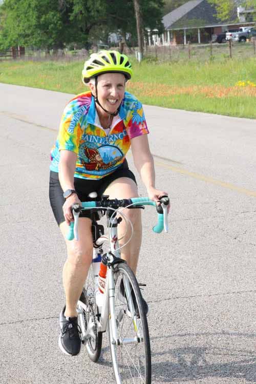 Bright female cyclist wearing a colorful jersey and yellow helmet riding on a rural road.
