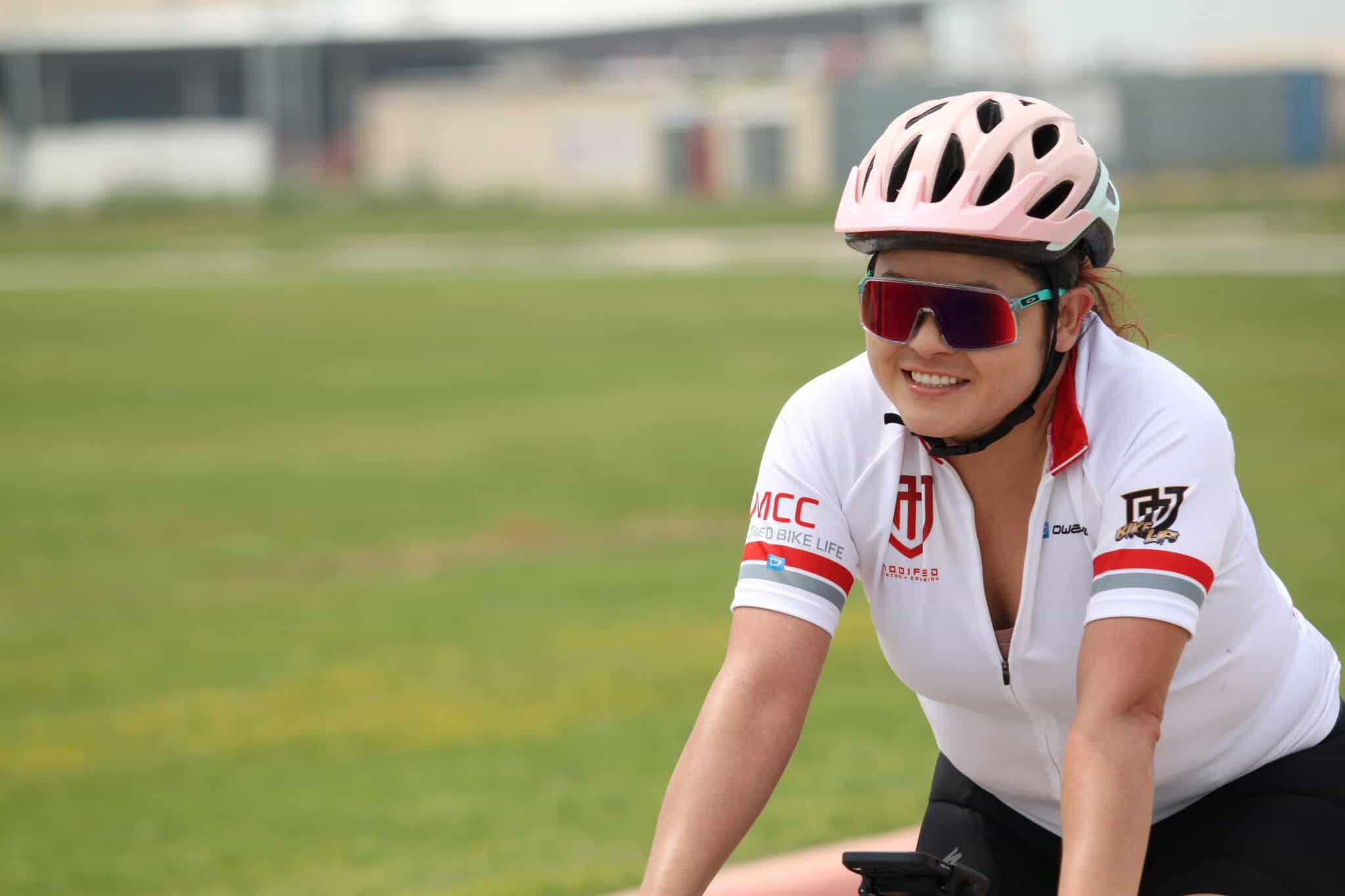 Smiling female cyclist riding outdoors on a sunny day, wearing a pink helmet and red sunglasses.