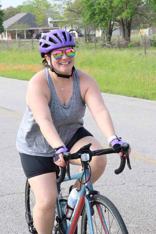 Bright smiling woman in purple helmet riding a bike in a scenic outdoor setting.