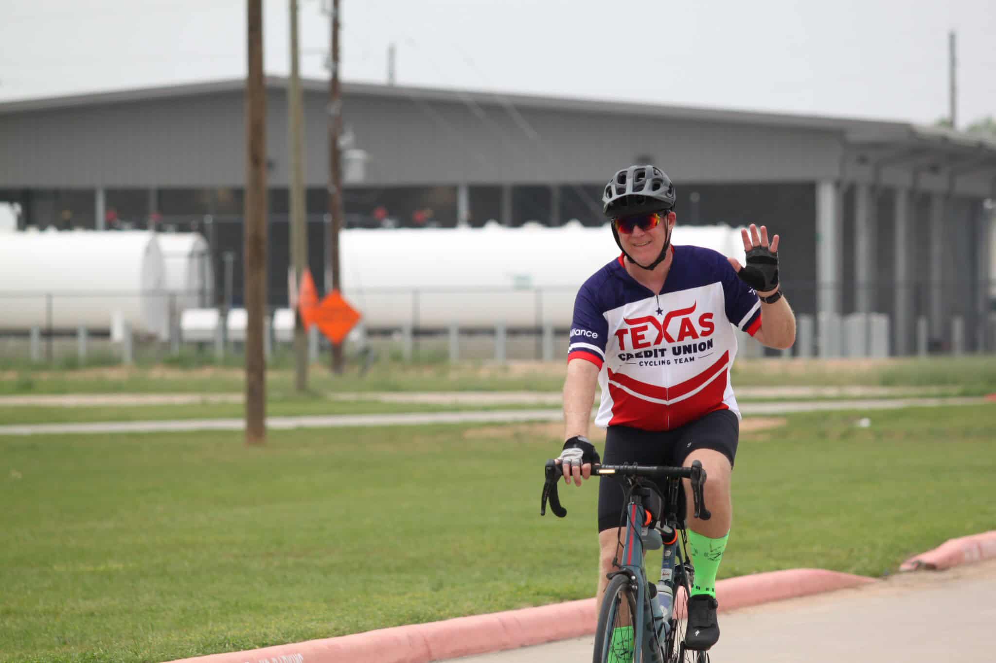 Smiling cyclist waving while riding on a bike trail, promoting Bluebonnet Express cycling event.