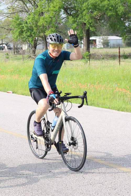 Smiling cyclist waving while riding a bike on a scenic road with green trees in the background.