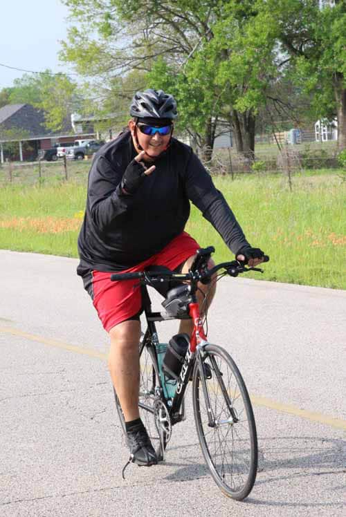 - Man riding a bicycle with a smile, wearing helmet and sunglasses on a sunny day.