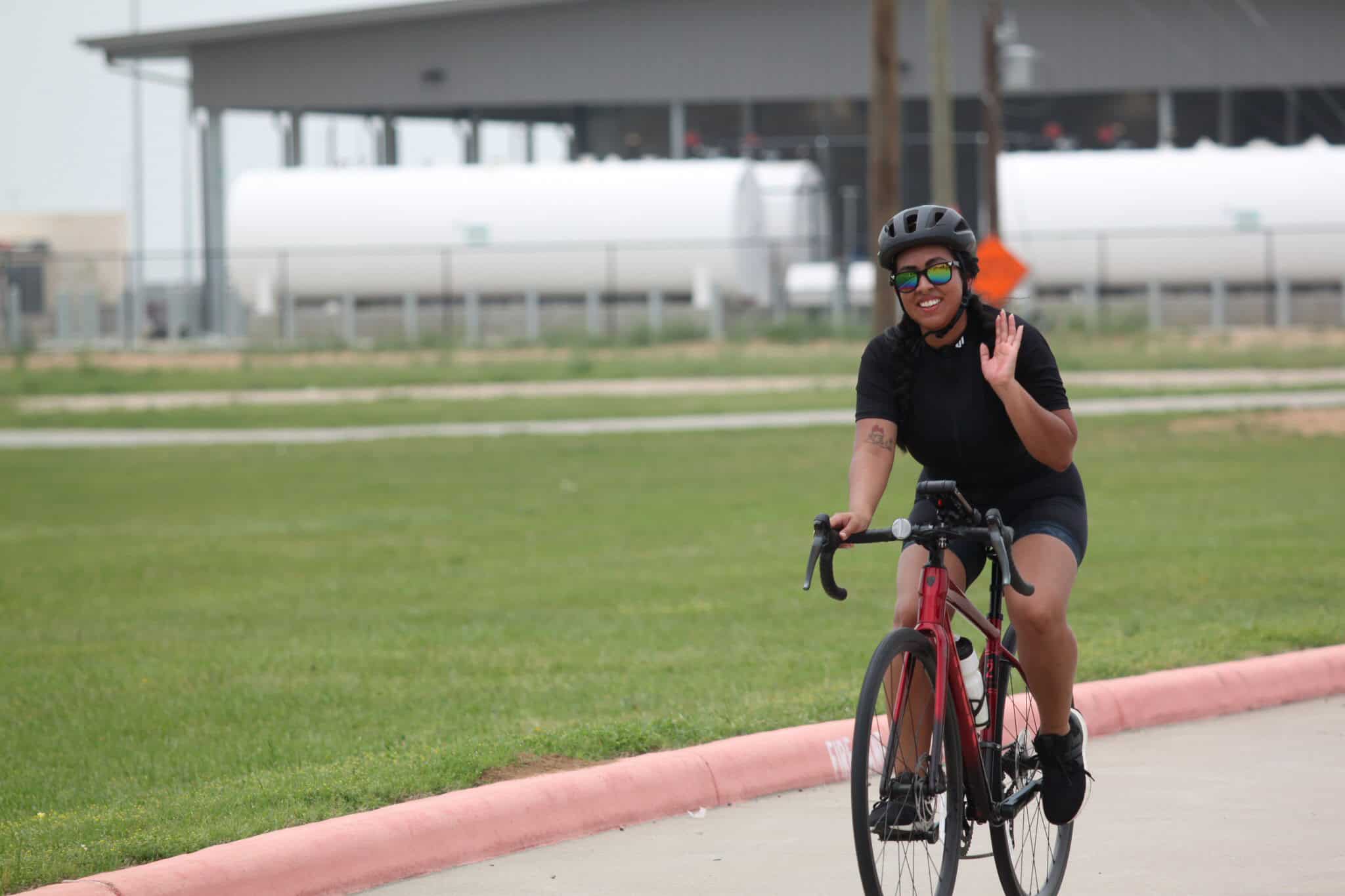 Bright woman riding Bluebonnet Express bike, smiling and waving at outdoor park scene.