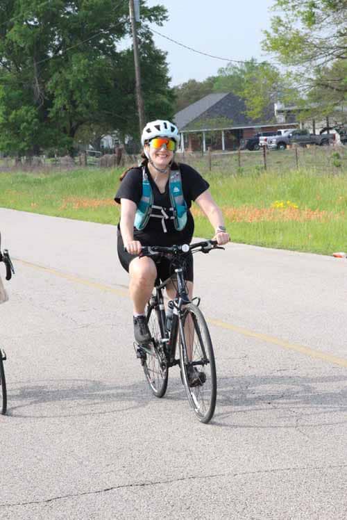 Bright cyclist wearing helmet and sunglasses riding a bike along a sunny rural road for Bluebonnet Express event.