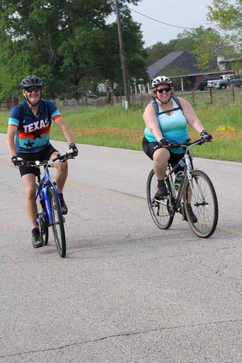 Riding bikes on outdoor road with greenery in background, two women enjoying cycling adventure with helmets and smiles.