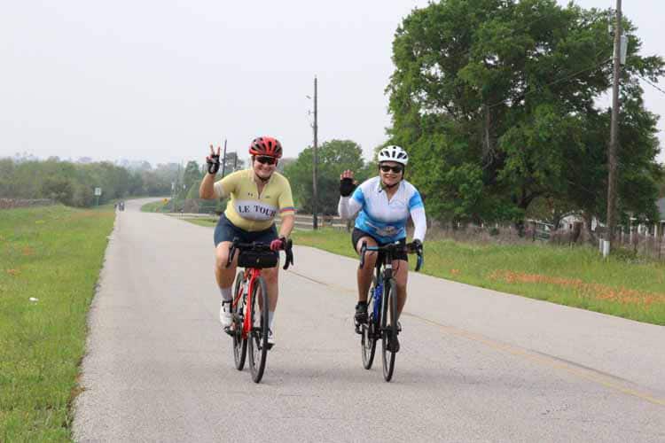 Brightly dressed cyclists riding on rural road with lush greenery and blue skies, promoting Bluebonnet Express bike tours.
