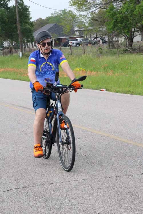 Bright blue cycling jersey with yellow accents, black helmet, and orange gloves on a road bike.