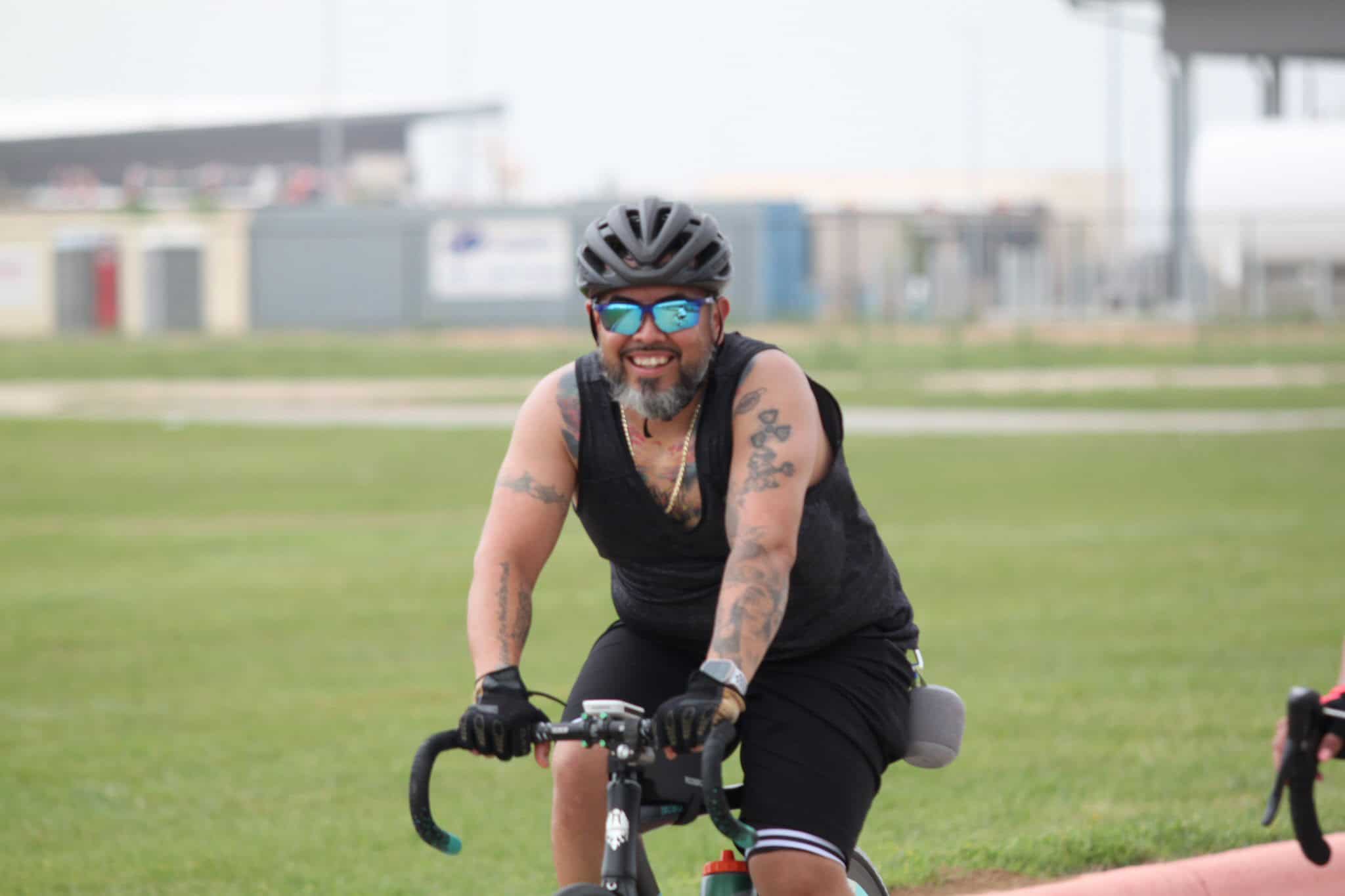 Smiling man riding a bicycle on a grassy field, wearing a helmet and sunglasses, enjoying outdoor biking fun.