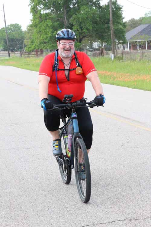 Smiling man cycling on a rural road with lush greenery and homes in the background, enjoying outdoor exercise.