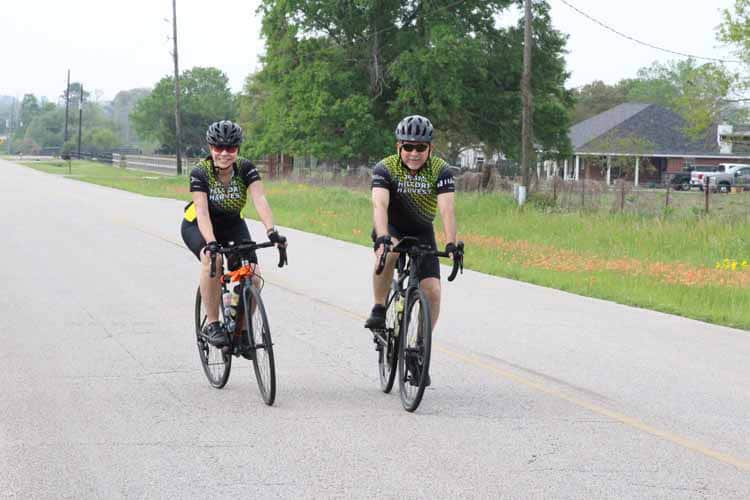 Bike riders enjoying a scenic ride on a peaceful road with green trees and houses in the background.