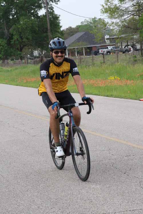 Man riding bicycle on street in cycling gear and helmet for Bluebonnet Express event.
