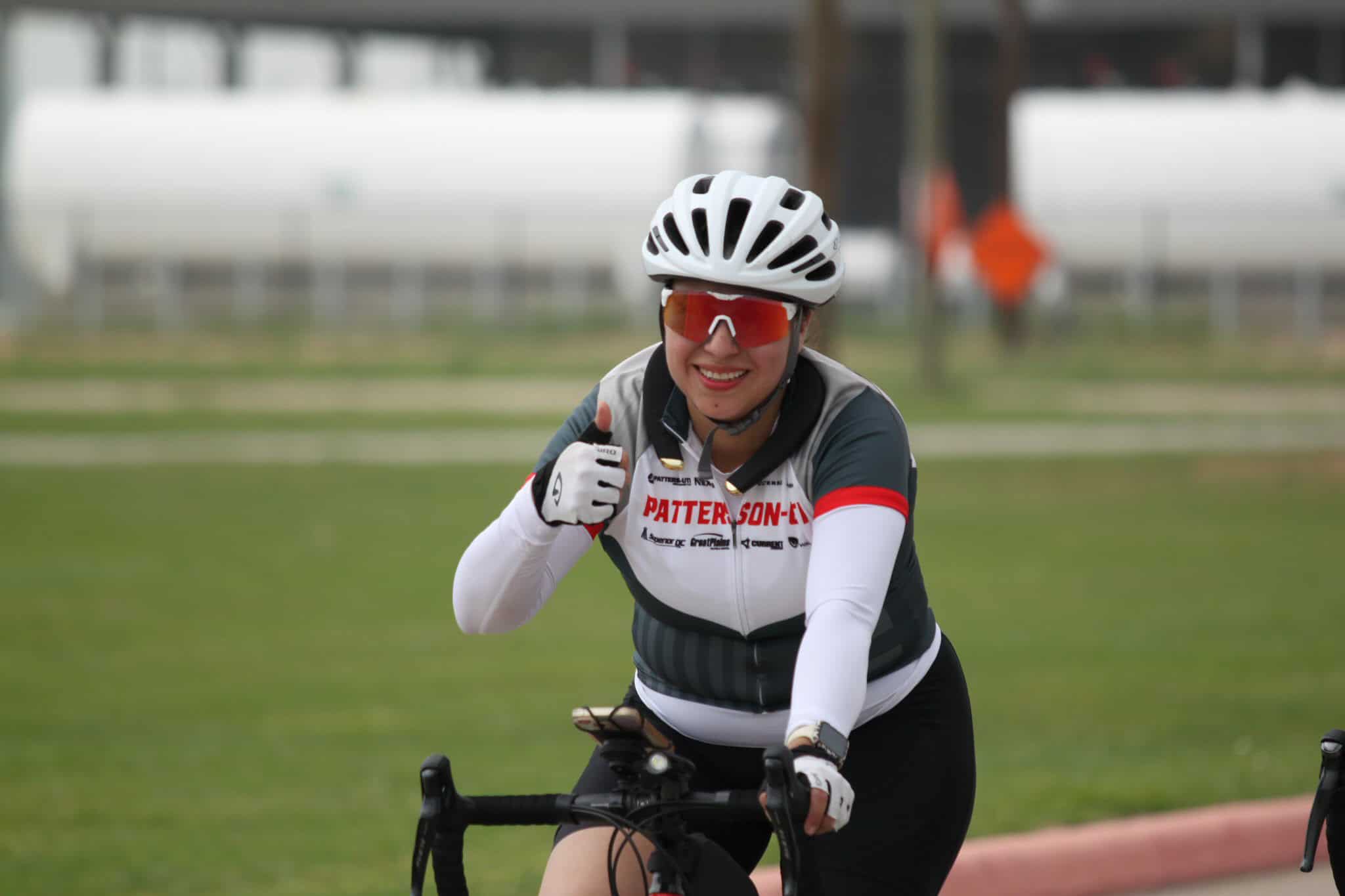 Bright female cyclist smiling with thumbs up, wearing helmet and sunglasses, riding bike outdoors.