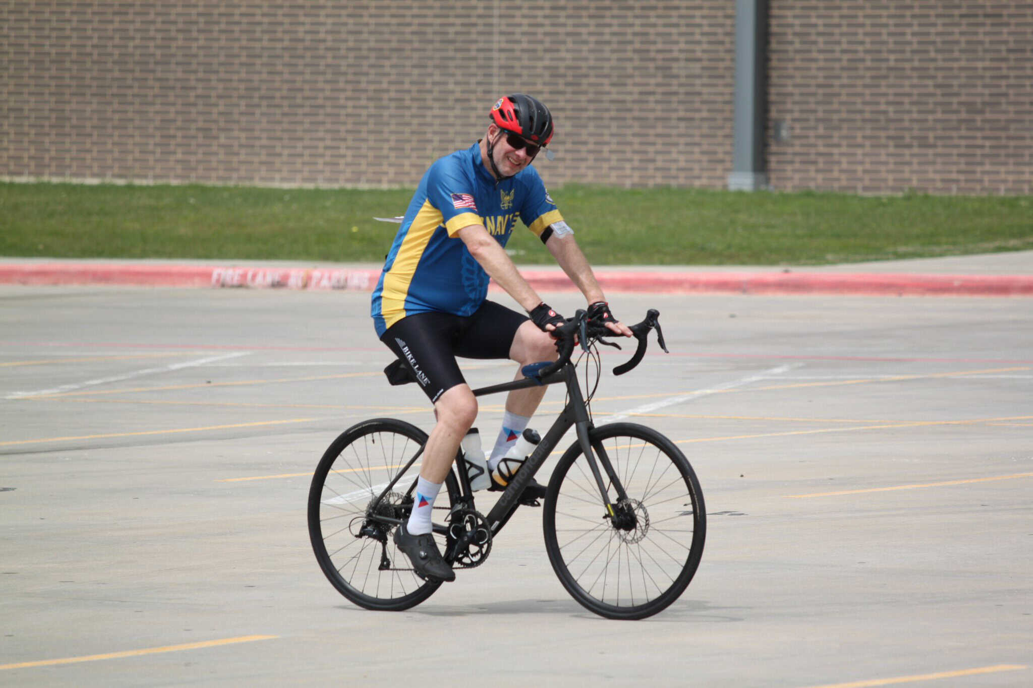 Smiling man riding a black Trek bicycle, wearing a blue and yellow Team Navy jersey, safety helmet, and sunglasses on an empty parking lot.