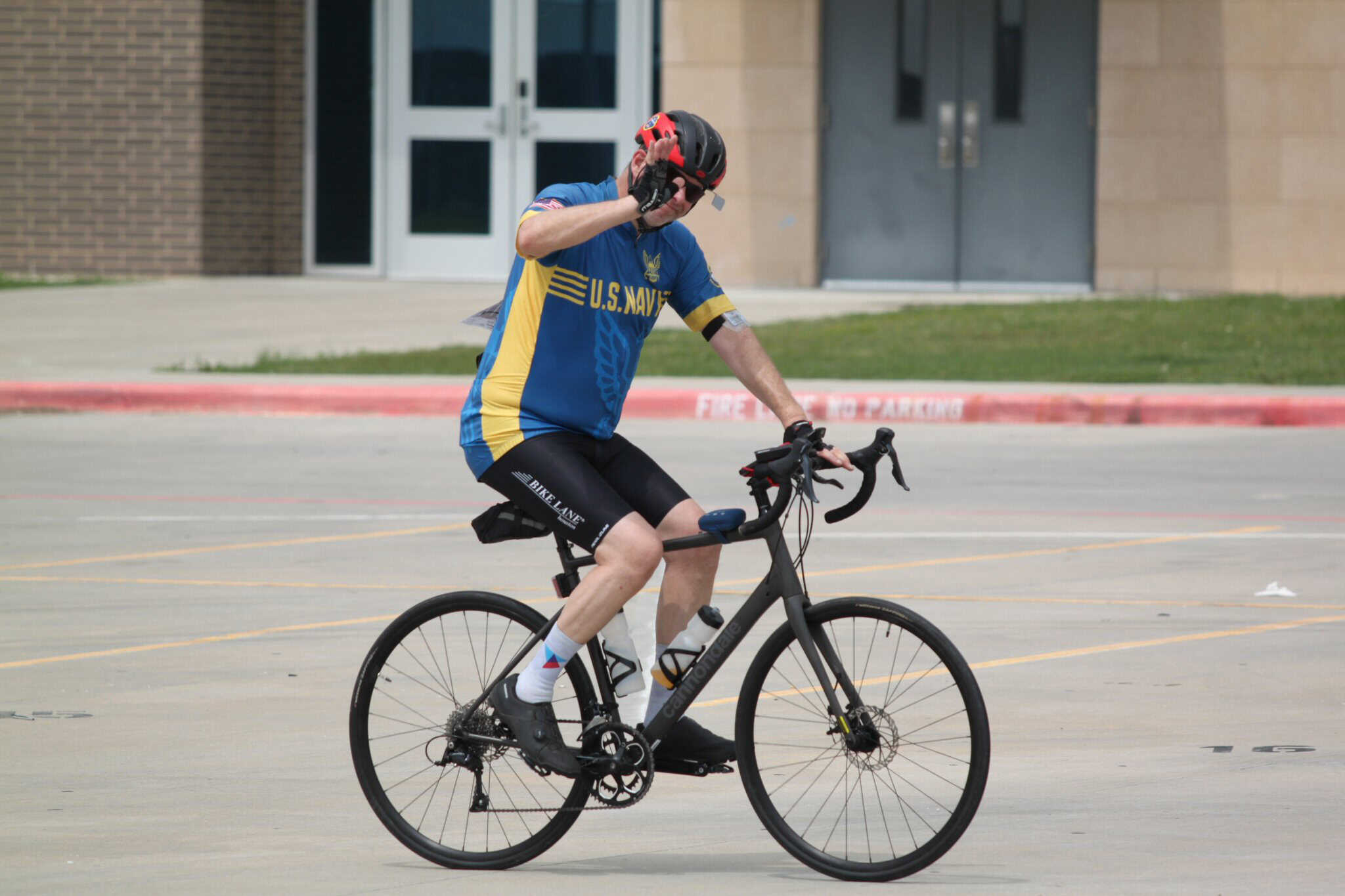 U.S. Navy cyclist on a black electric bike in a parking lot, wearing a helmet and sports gear.