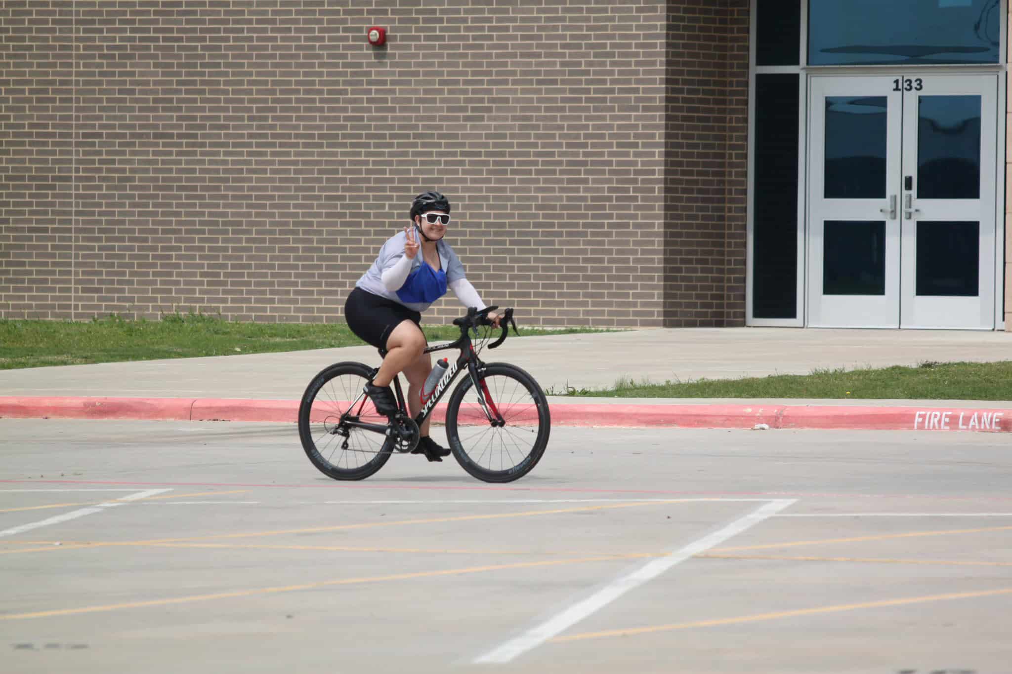 Bright woman riding a bike outside wearing a helmet and sunglasses, making peace sign gesture.