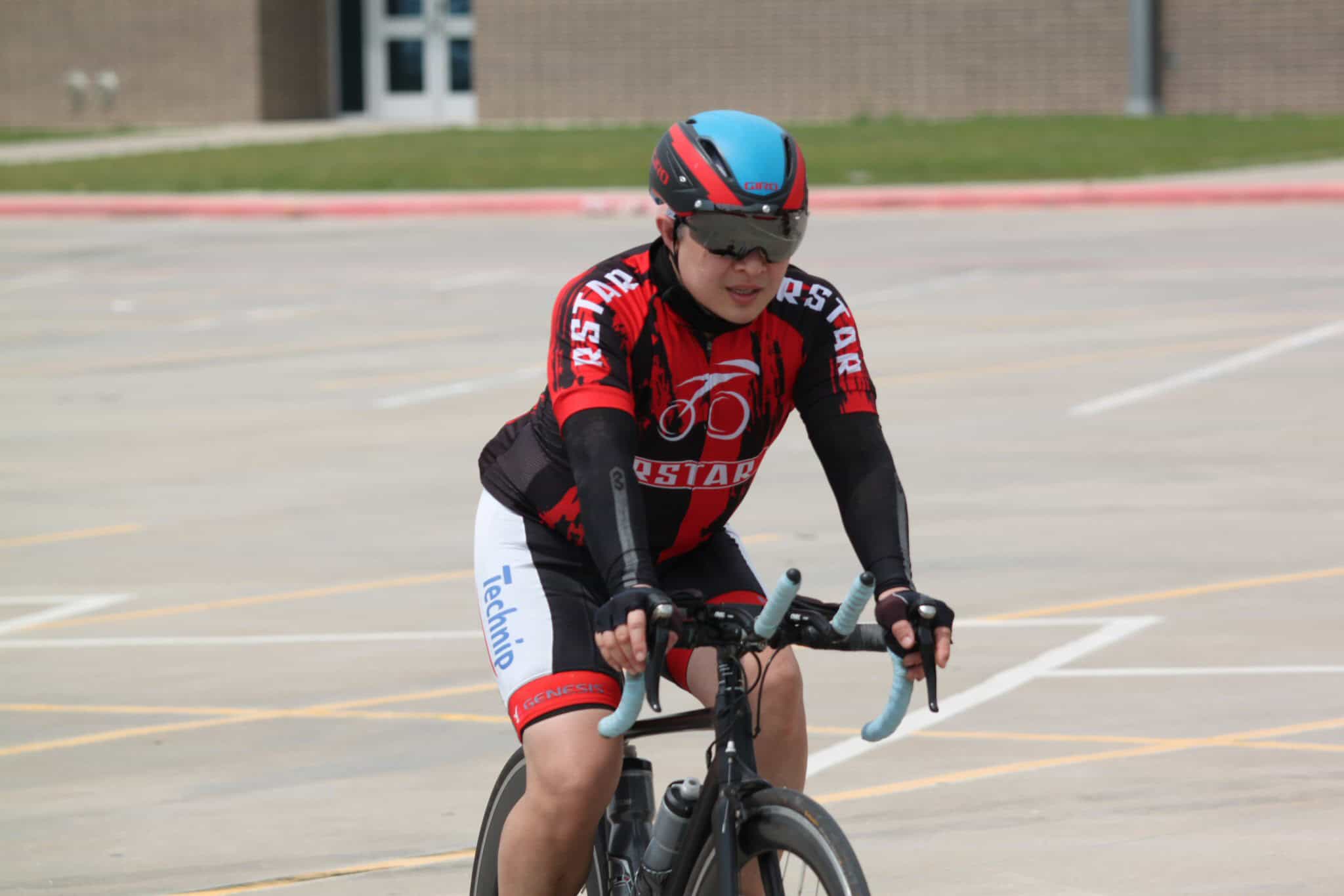 Bright cyclist wearing a red and black jersey riding a bike on a parking lot.