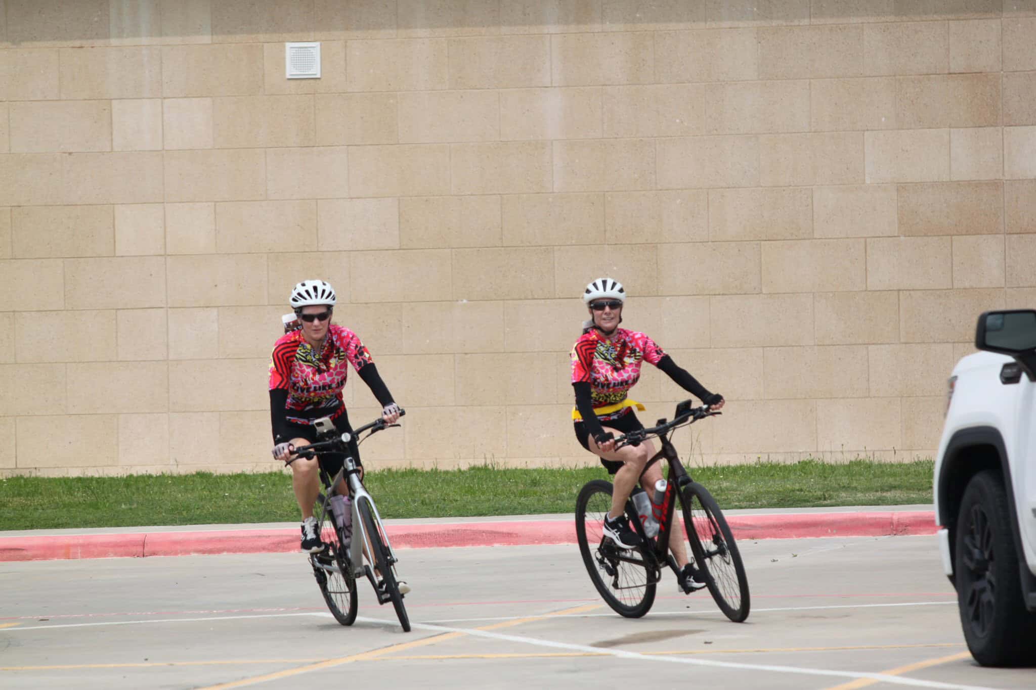 Bright cyclists in colorful jerseys ride bikes in a parking lot on a sunny day.