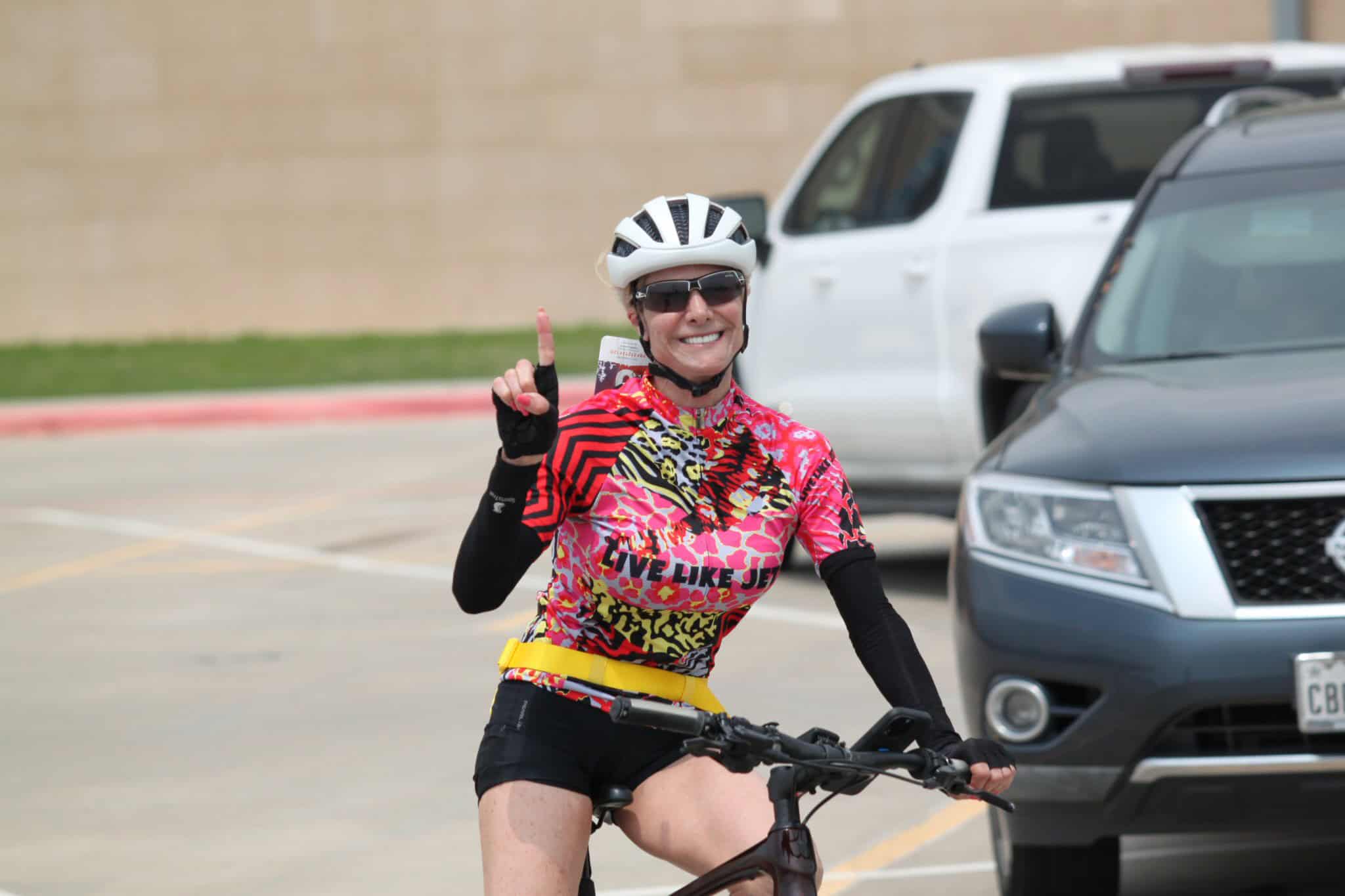 Bright female cyclist wearing a colorful jersey, helmet, and sunglasses, smiling and pointing up in a parking lot.