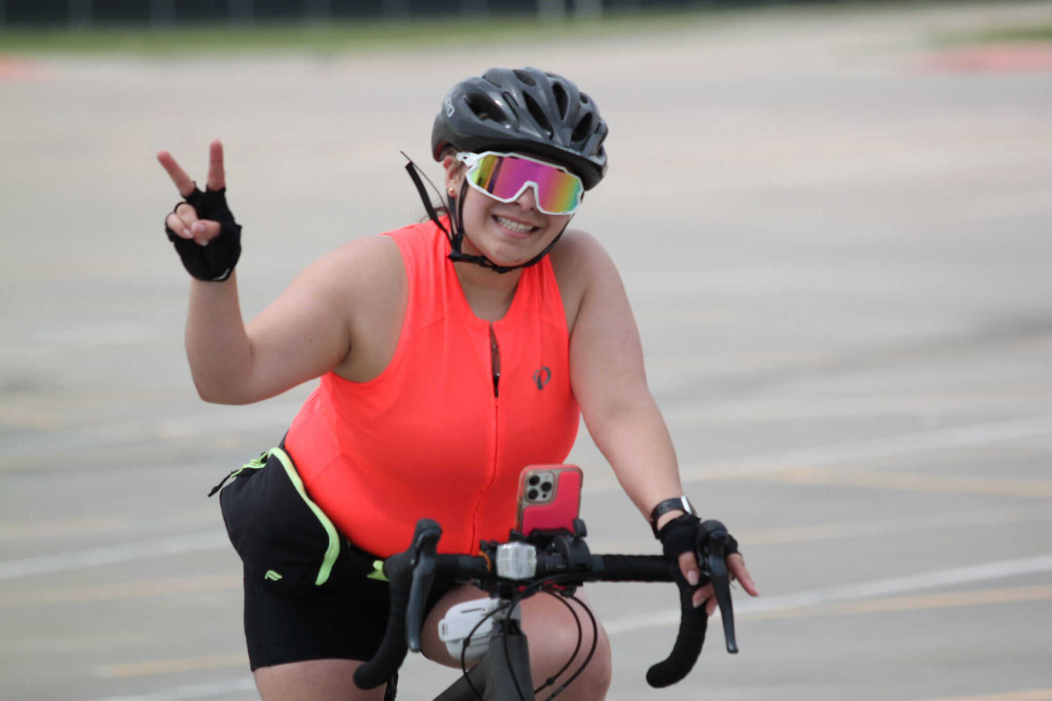 Vibrant female cyclist smiling and making a peace sign while riding her bike in a parking lot.