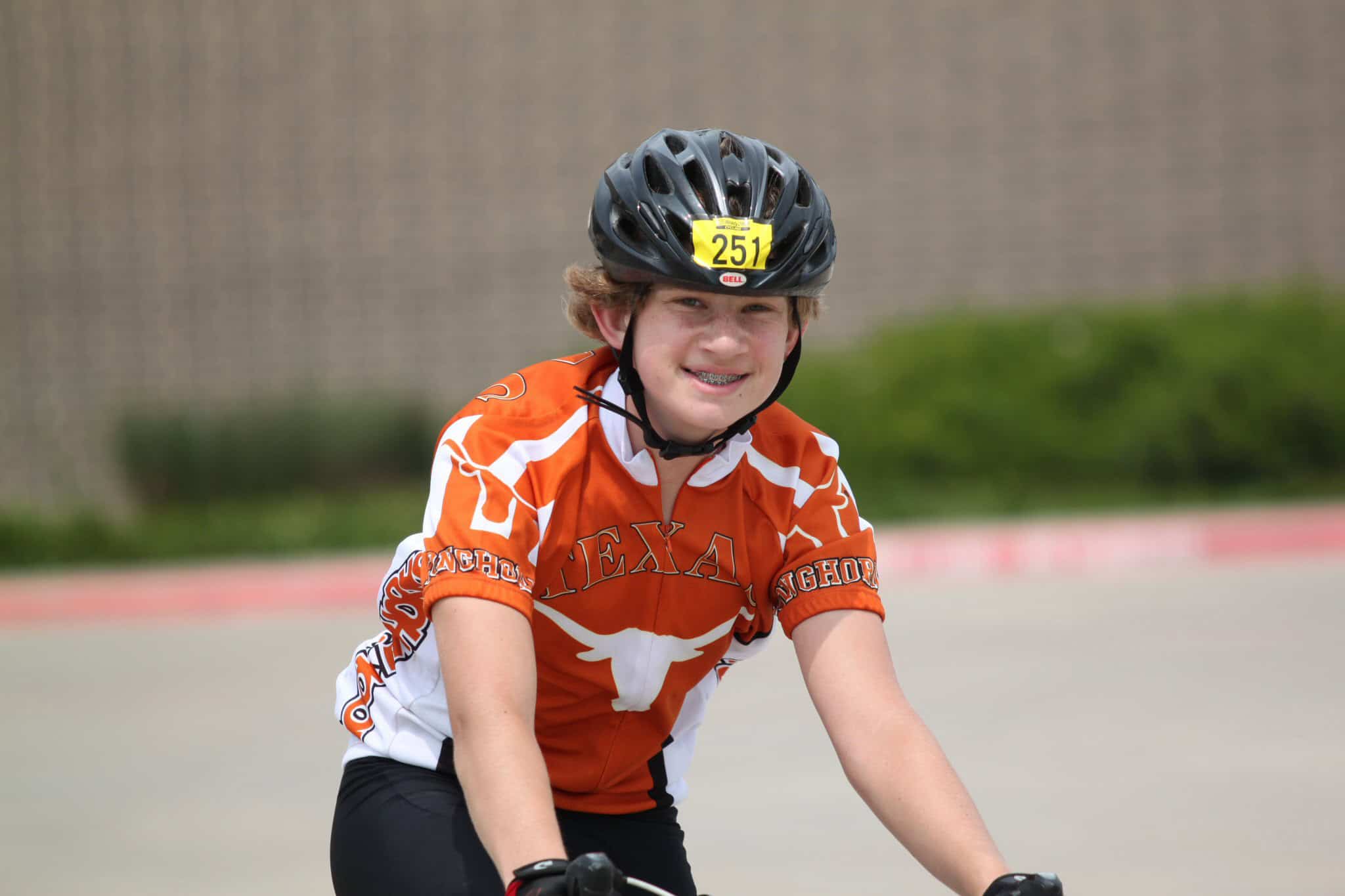 Young cyclist wearing a Texas Longhorns jersey and helmet riding a bike outdoors.