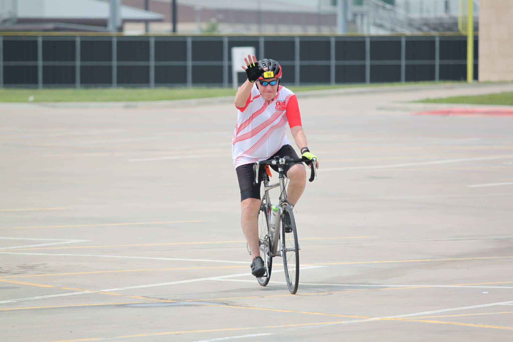 Cyclist waving while riding on an empty parking lot, promoting Bluebonnet Express bike event.