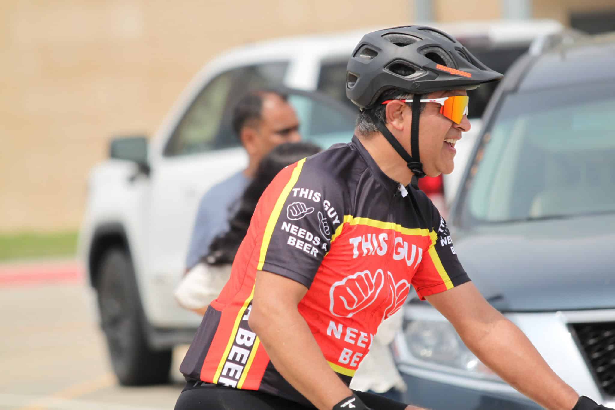 Vibrant cyclist wearing helmet and sunglasses with "This Guy Needs Beer" jersey during Bluebonnet Express event.