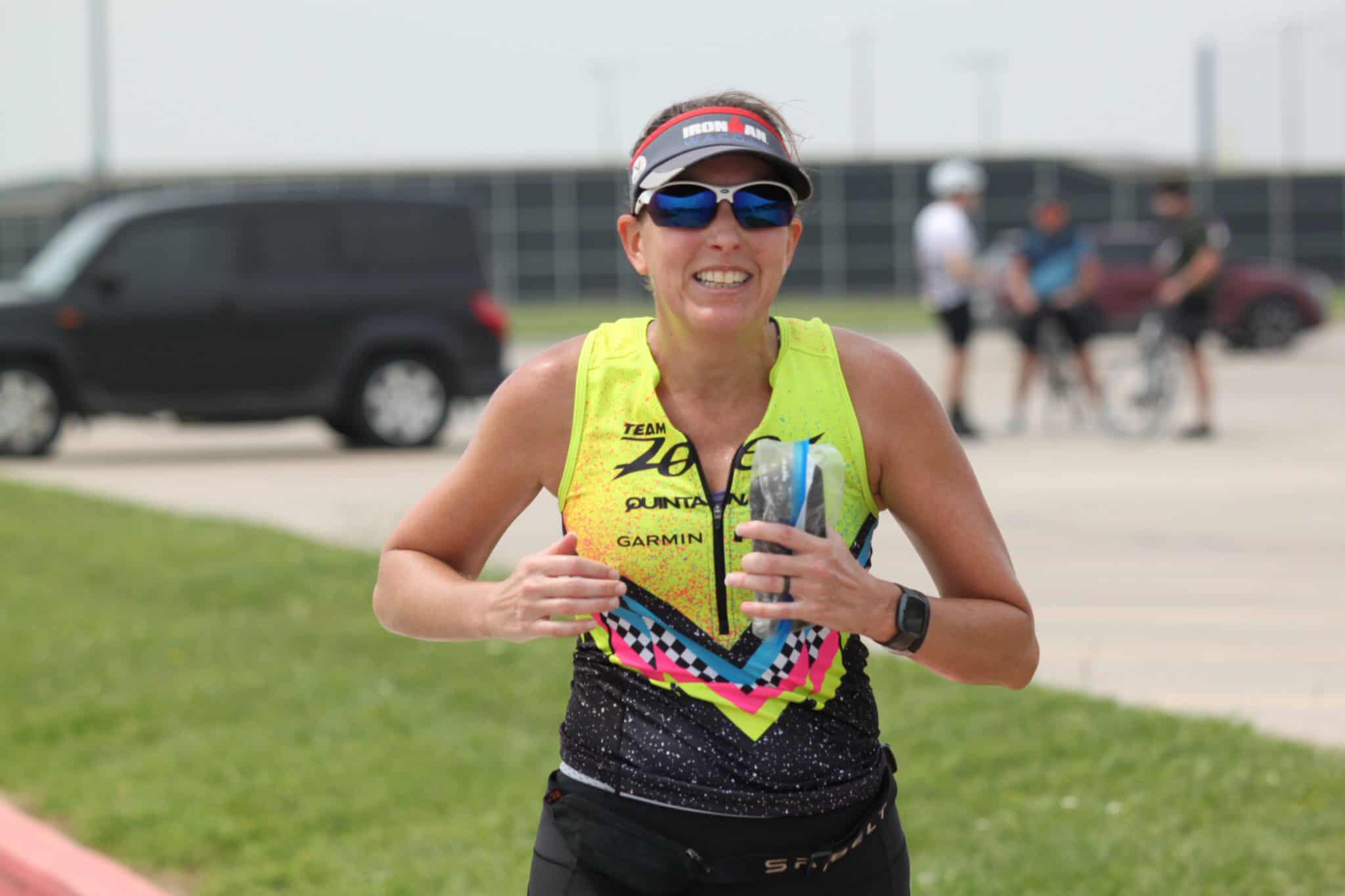 Vibrant female triathlete smiling with hydration, wearing bright race gear and sunglasses, during a race event.