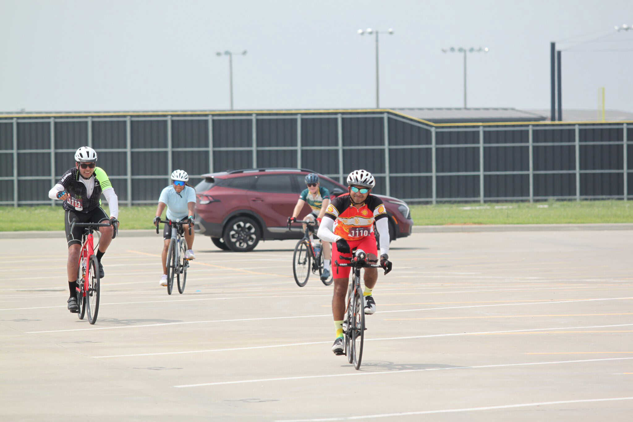Brightly dressed cyclists riding outdoors during daytime in a parking lot with a gray fence in the background.