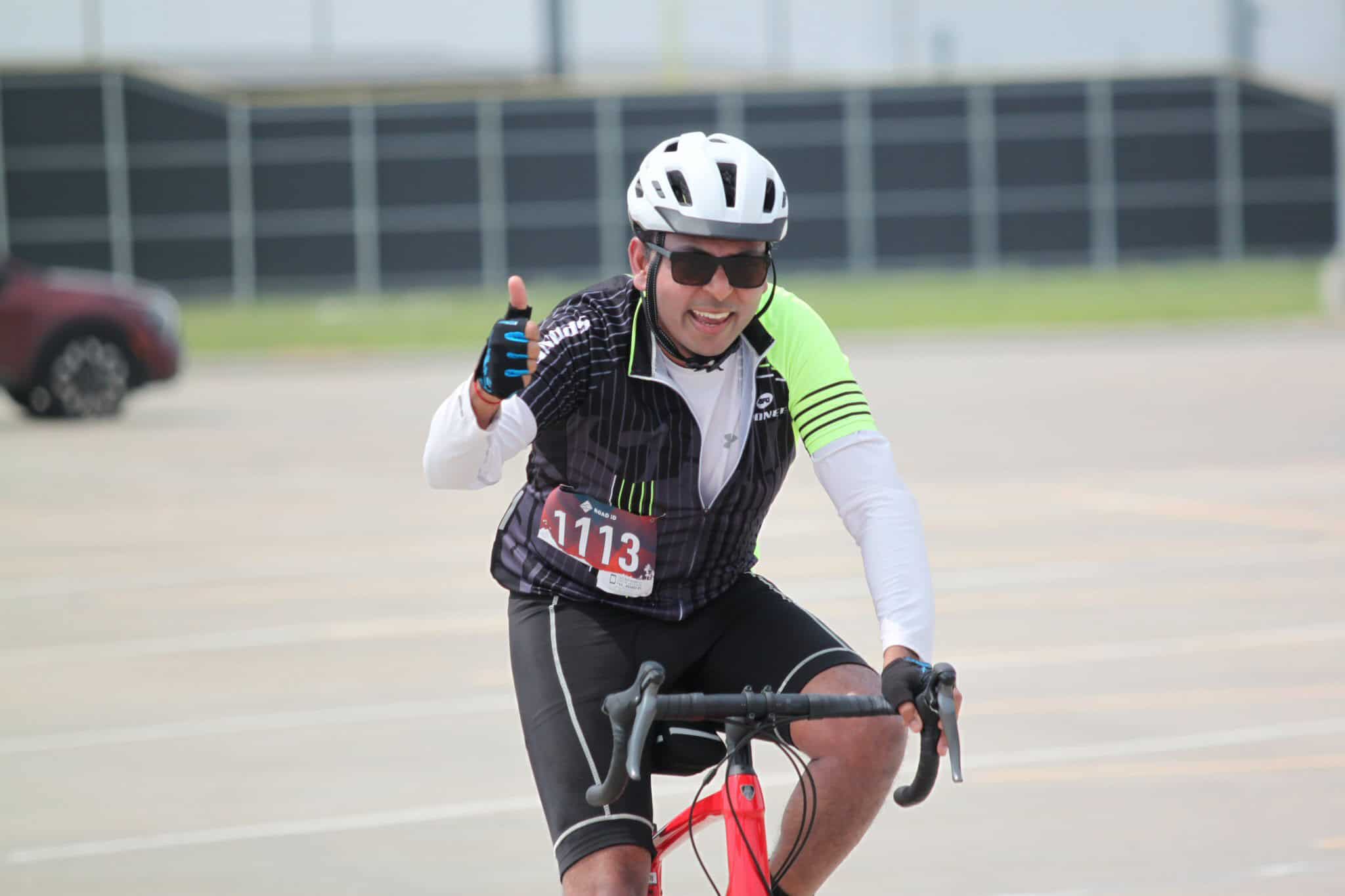 Happy man riding a bicycle, wearing cycling gear, helmet, sunglasses, and giving a thumbs-up at a cycling event.