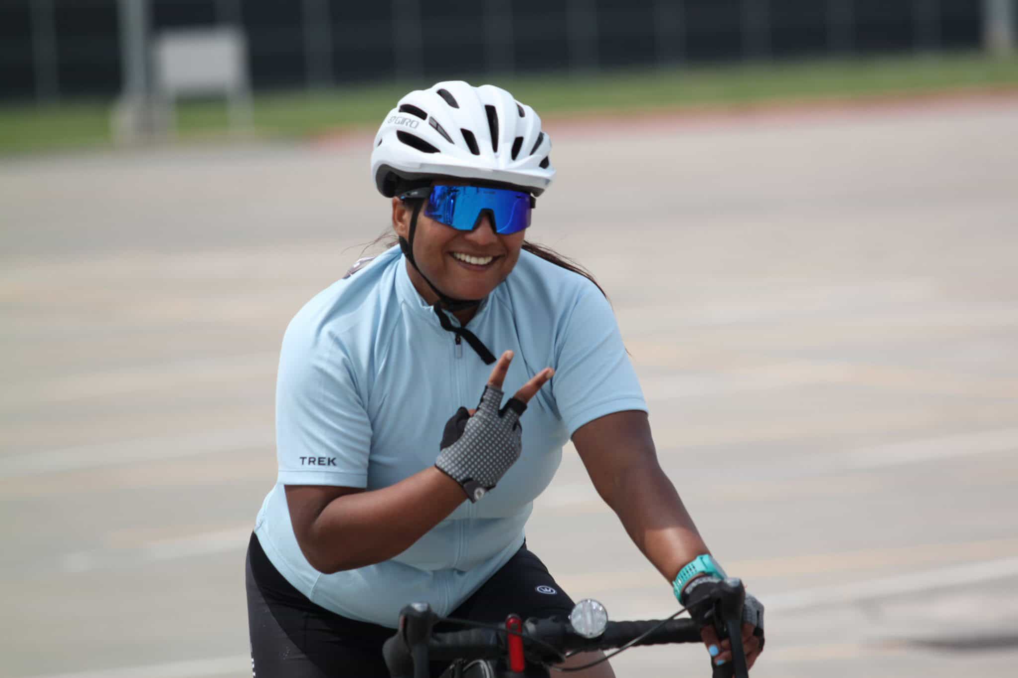 Smiling woman cyclist with helmet and sunglasses making a peace sign during outdoor ride.