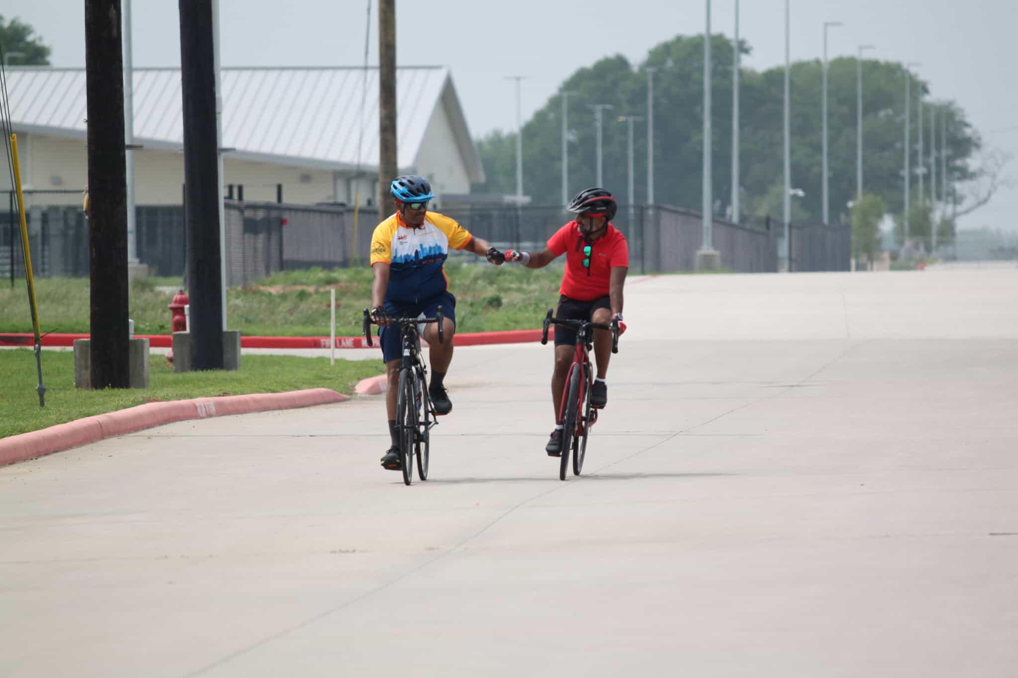 Two cyclists sharing a handshake while riding bikes on a paved trail, emphasizing community, health, and cycling in Bluebonnet Express area.
