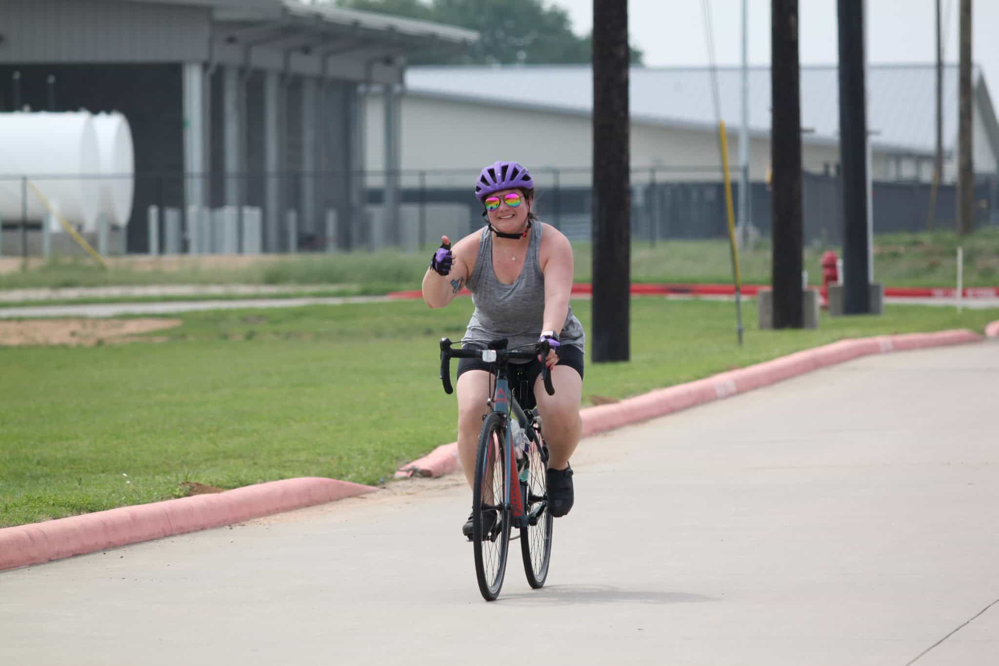 Bright smiling woman riding a bicycle on a paved path, wearing a helmet and sunglasses, enjoying Bluebonnet Express cycling event.