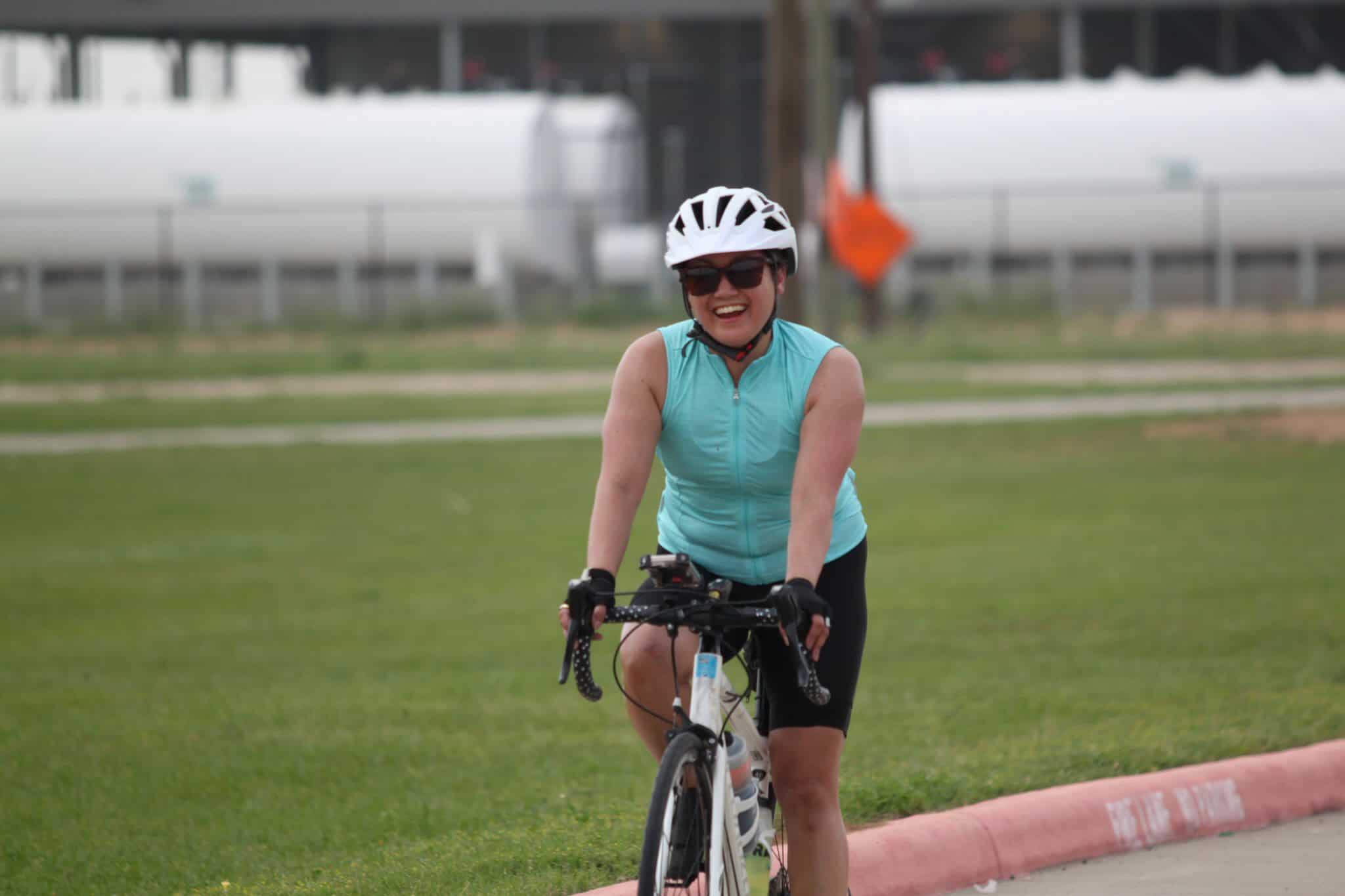 Bright woman cyclist smiling, wearing helmet and sunglasses on a green field, promoting Bluebonnet Express bike services.