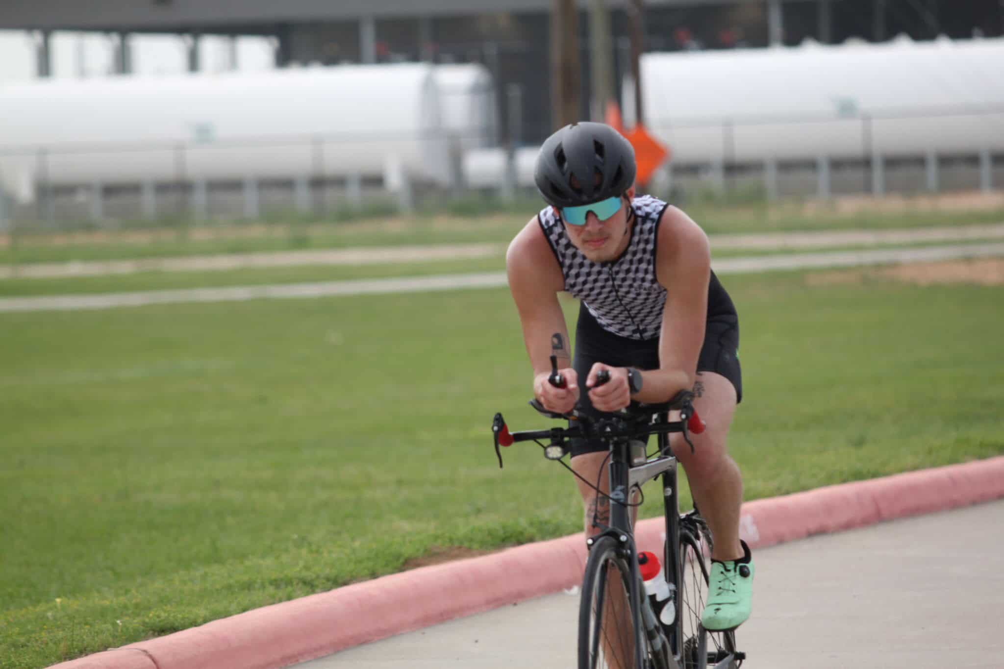 High-performance cyclist wearing helmet and sunglasses riding a black road bike on a paved path.