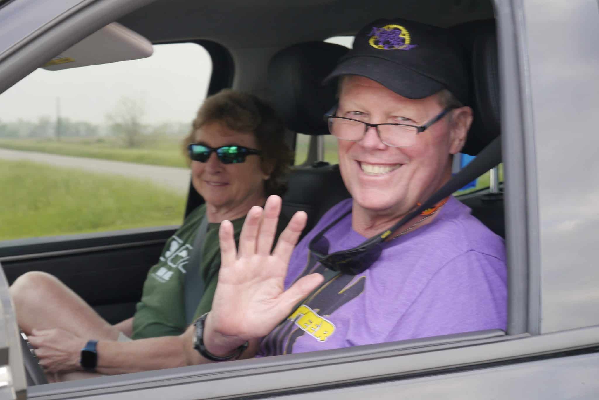 Smiling man waving from car window at Bluebonnet Express event, with companion on road trip.