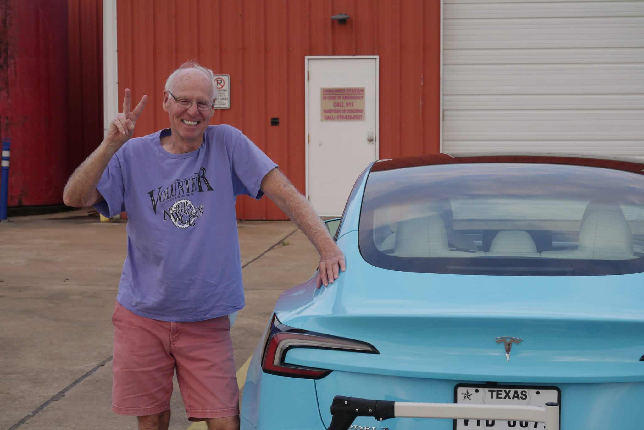 Bright smiling man making peace sign next to a custom blue Tesla car at Bluebonnet Express.