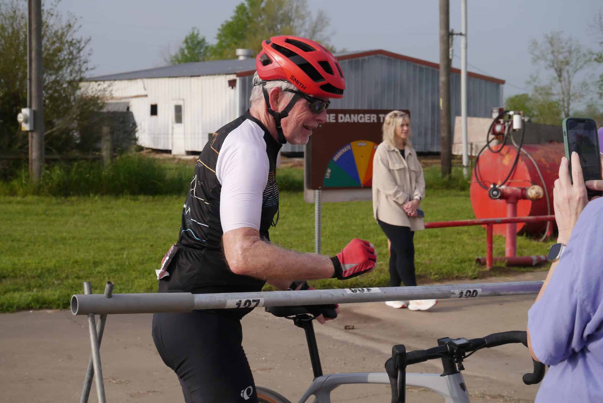 Older man in cycling gear and red helmet smiling during a bike race.
