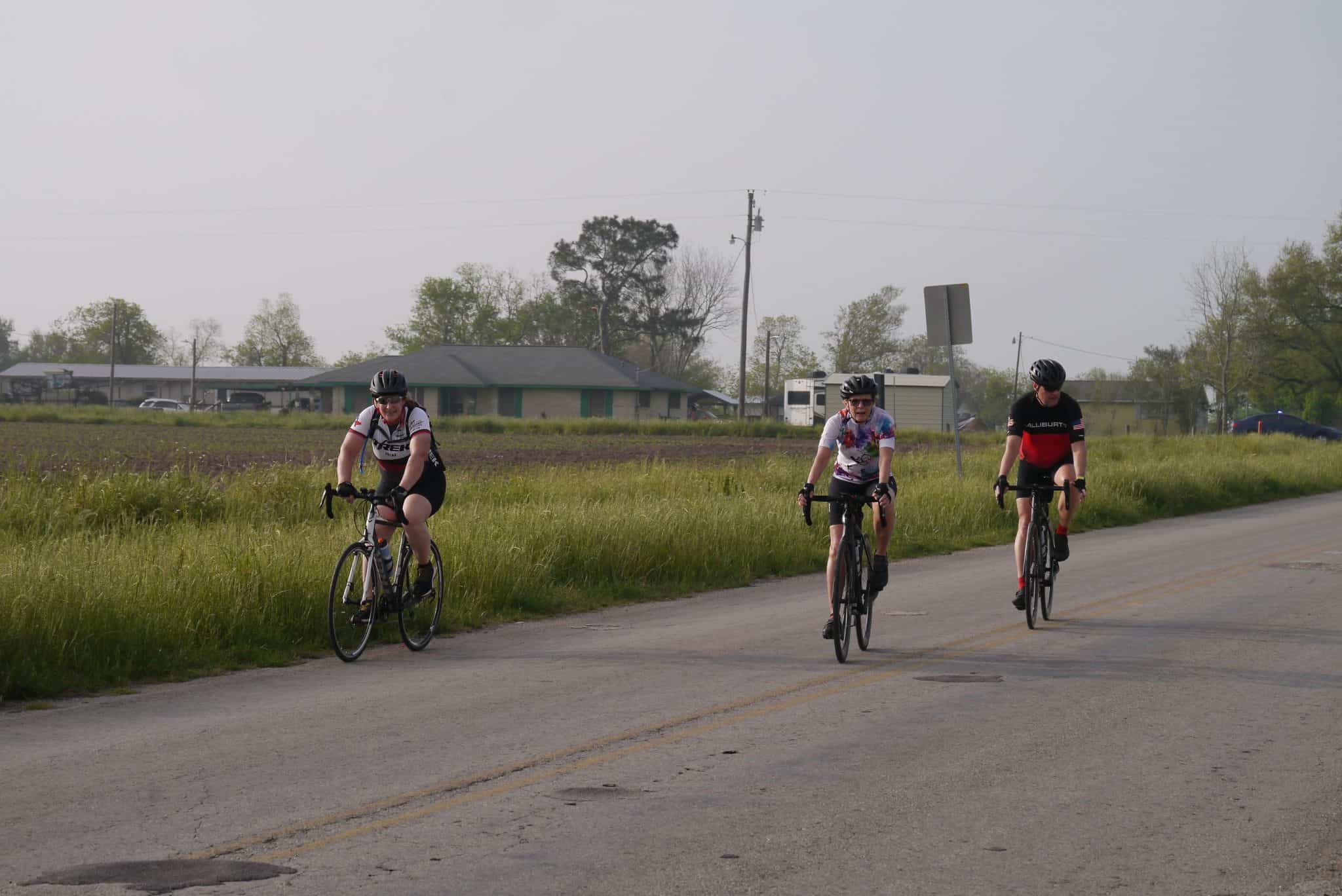 Cyclists riding on a rural road during a bike event with grassy fields and houses in the background.